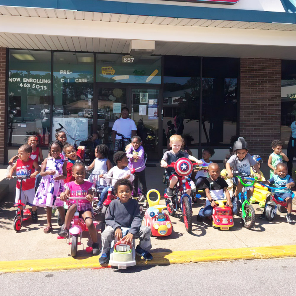 A group of children poses on bicycle and ride-ons outside a building, enjoying their day together.