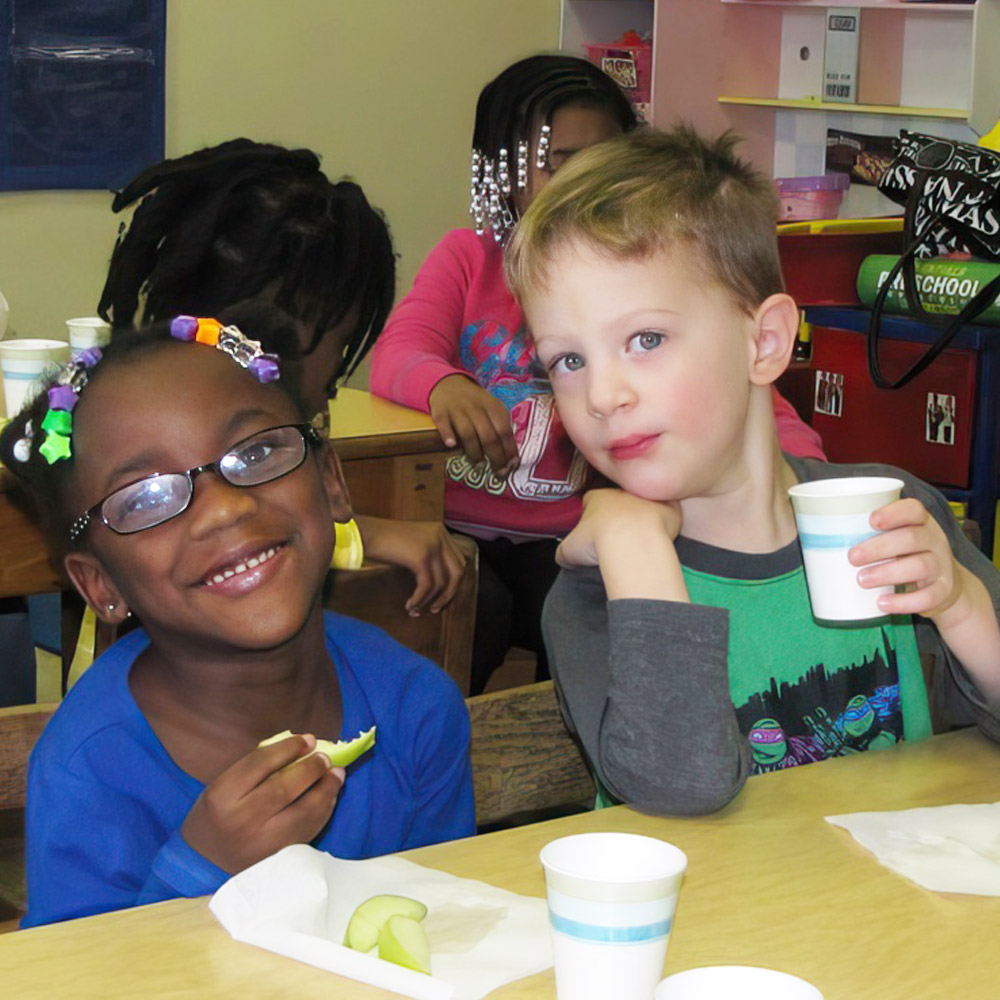 Two children smile while enjoying snacks, one holding a slice of apple and the other with a cup.