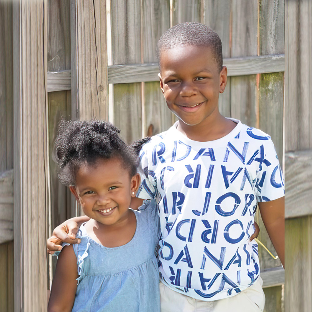 Two smiling children pose together outdoors, showcasing friendship against a wooden backdrop.