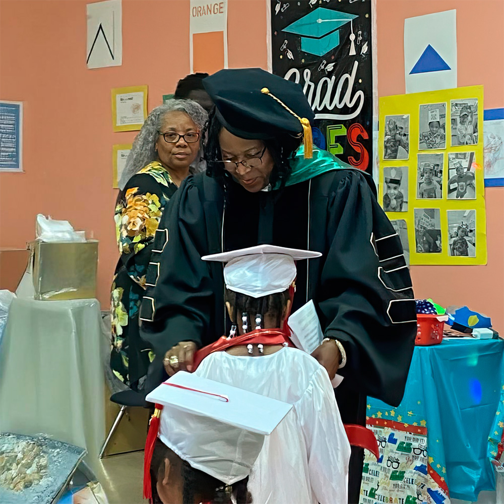 A teacher in graduation attire adjusts the cap of a young graduate, celebrating a special milestone in a festive setting.