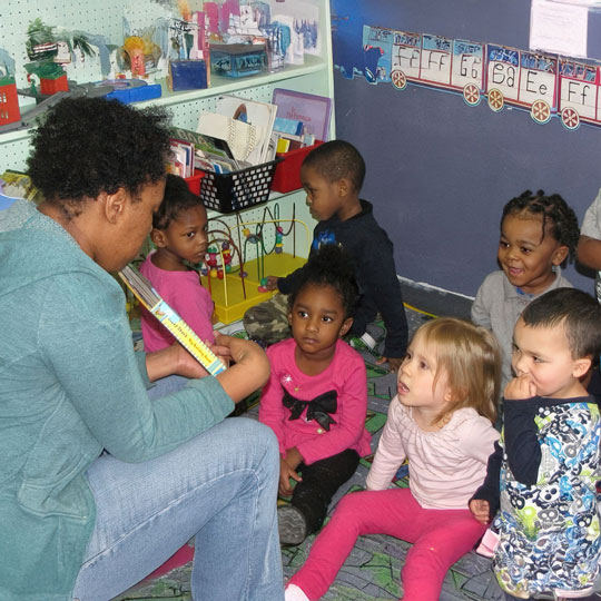 A teacher reads to a group of attentive children sitting on the floor, engaged in the storytelling session.